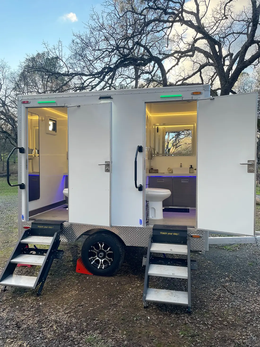 Two-door restroom trailer with steps parked under trees